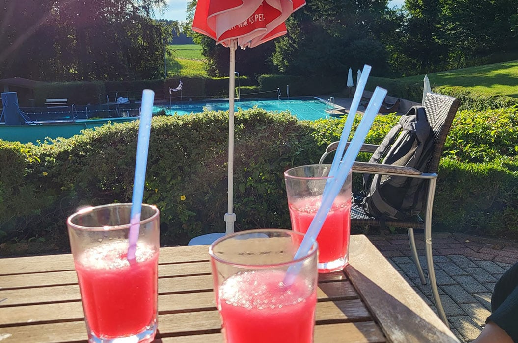 Drei Gläser rosa Getränke mit Strohhalmen auf Holztisch, im Hintergrund Pool und Sonnenschirm.